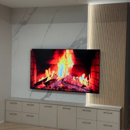 USŁUGI REMONTOWO-WYKOŃCZENIOWE DANIEL TĘCZA - Nowoczesna ściana TV z marmurowym panelem, drewnianymi lamelami z podświetleniem LED i szafką RTV w odcieniach beżu. Parkiet w jodełkę na podłodze.