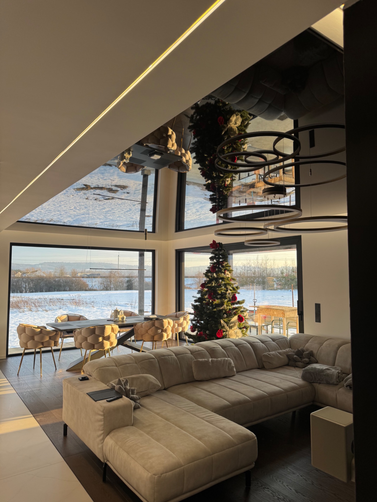 Nowoczesny salon z sufitem lustrzanym i choinką w Górze Motycznej. Duże okna z widokiem na zimowy krajobraz. Designerskie lampy i beżowa sofa.