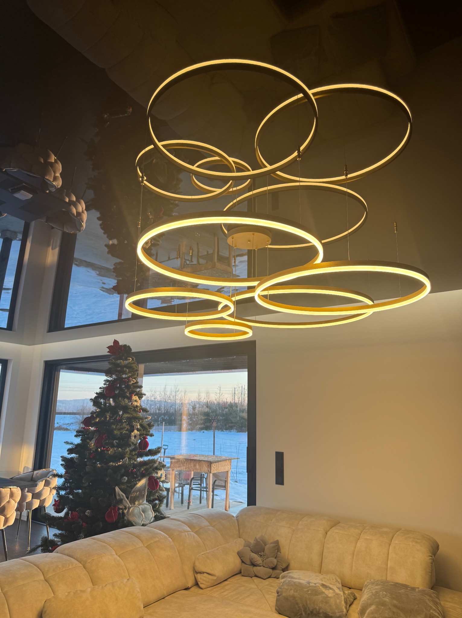 Nowoczesny salon z sufitem lustrzanym, oświetlony wiszącą lampą z okręgów LED. W tle okno z widokiem na zimowy krajobraz i choinka.