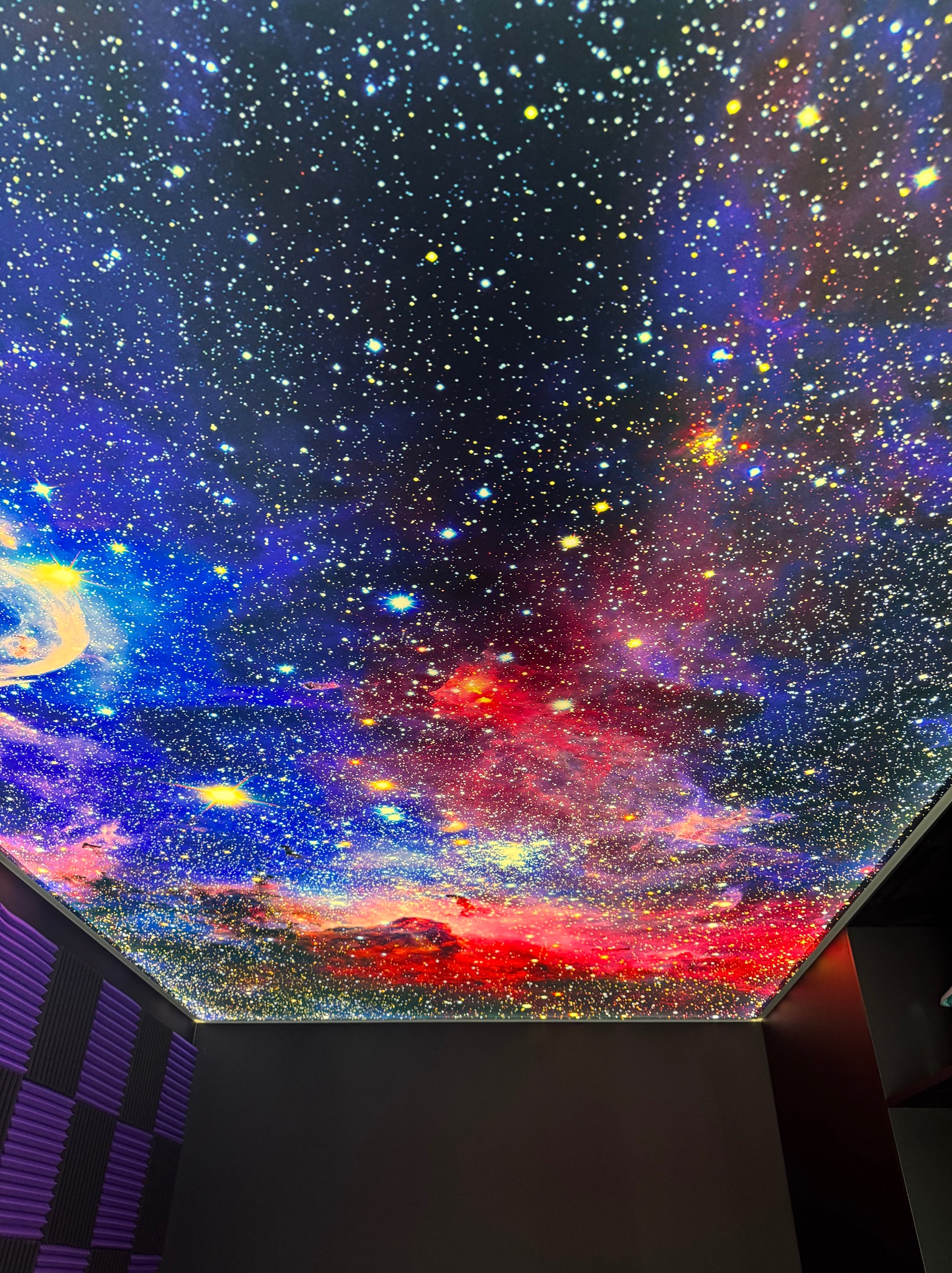 Sufit napinany z motywem kosmosu, widok z dołu. Efektowna instalacja w pomieszczeniu z panelami akustycznymi. Intensywne kolory i detale gwiazd.