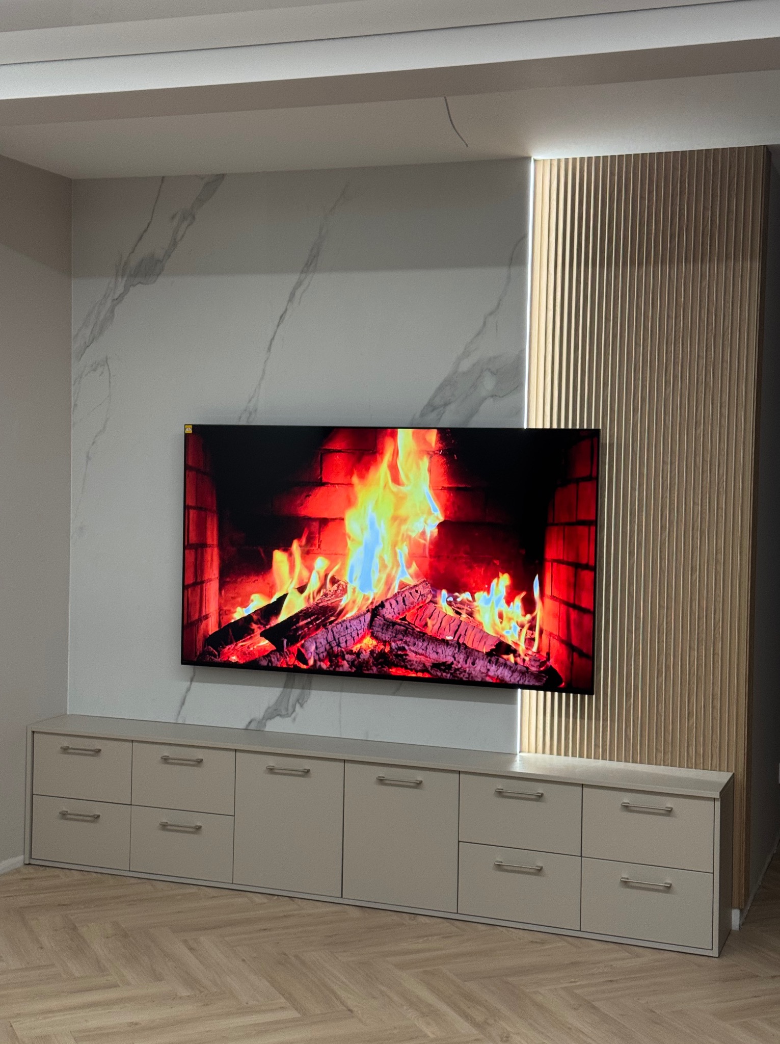 Nowoczesna ściana TV z marmurowym panelem, drewnianymi lamelami z podświetleniem LED i szafką RTV w odcieniach beżu. Parkiet w jodełkę na podłodze.