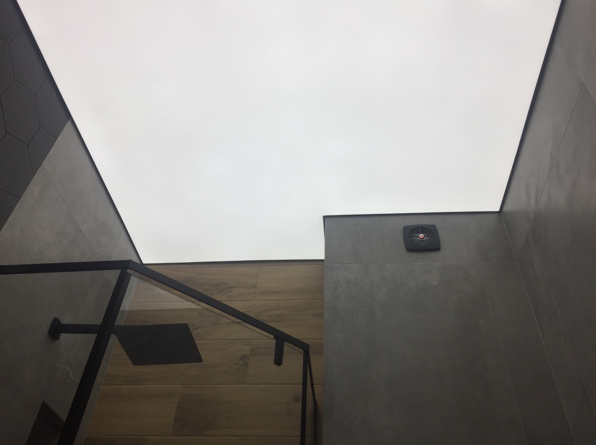 Nowoczesna łazienka z sufitem napinanym, szarą armaturą i drewnopodobnymi płytkami. Widoczny fragment kabiny prysznicowej i wentylator sufitowy.