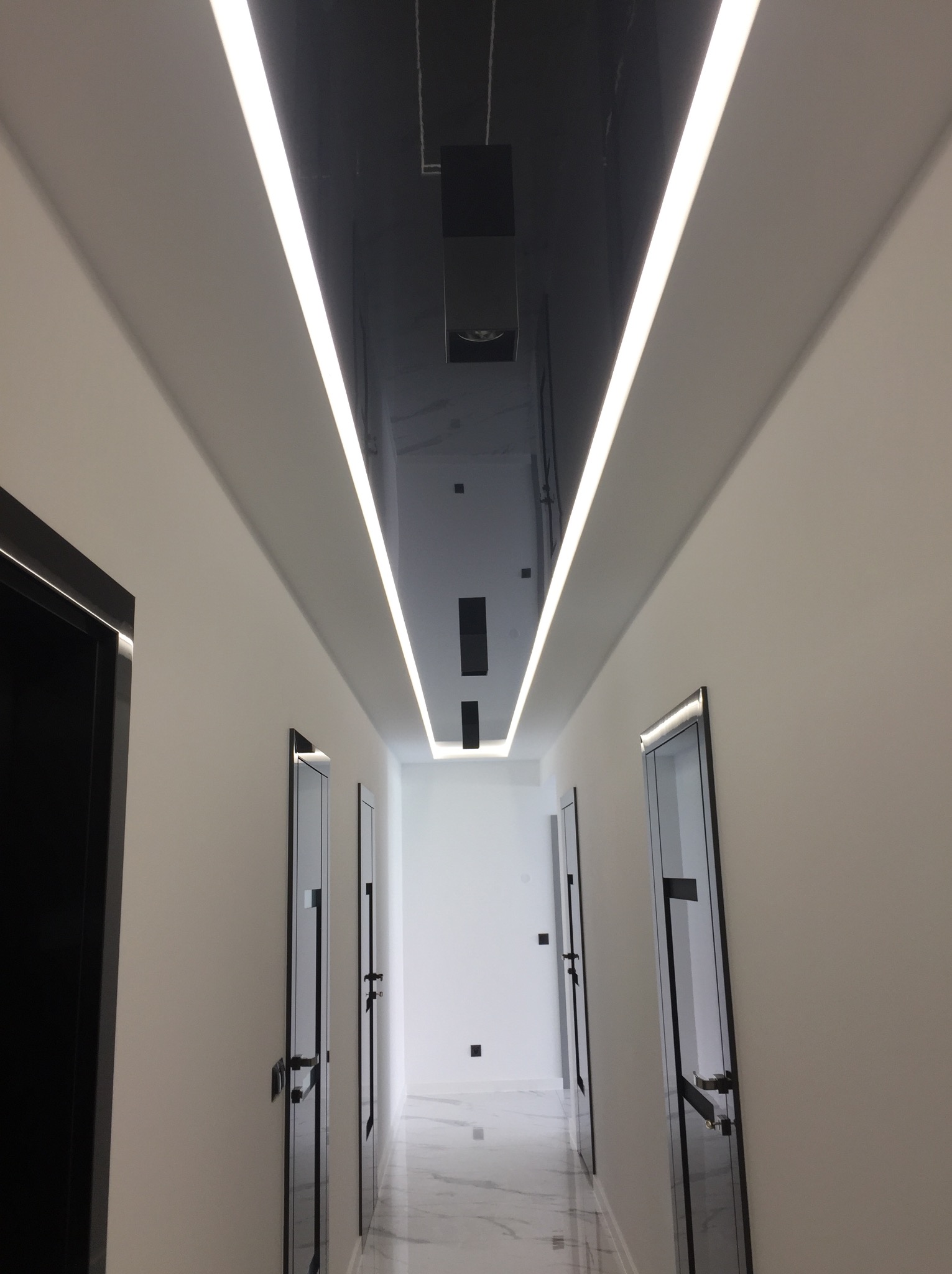 Błyszczący, czarny sufit napinany z oświetleniem LED wzdłuż korytarza z białymi ścianami i czarnymi drzwiami. Marmurowa podłoga odbija światło.