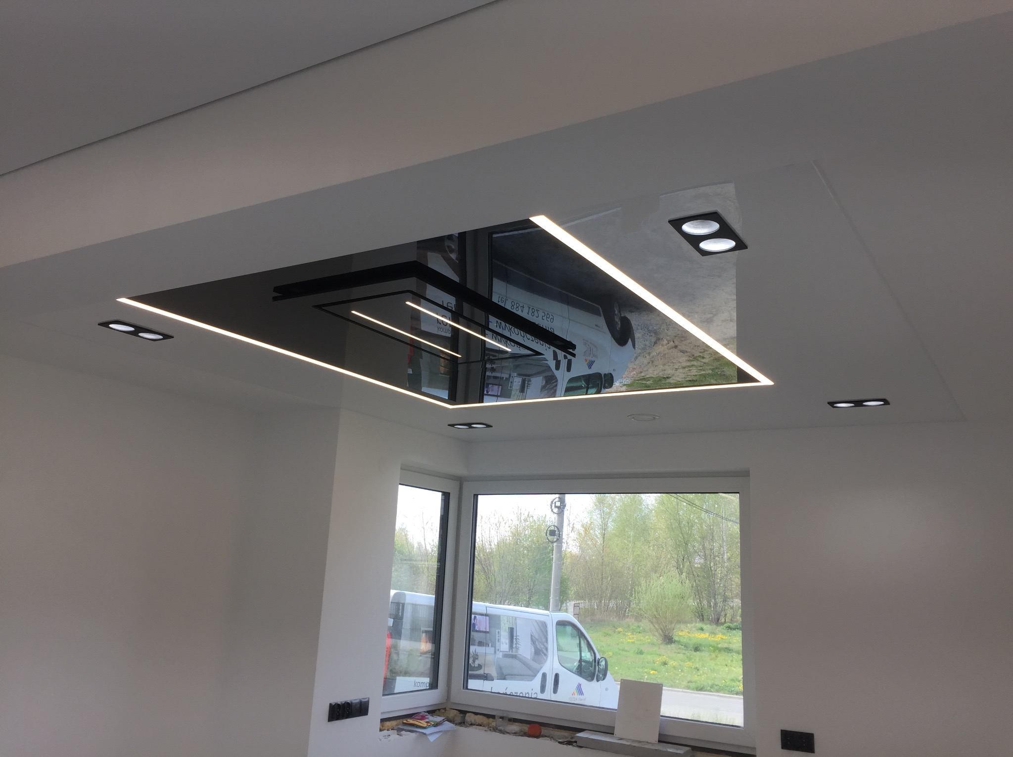 Nowoczesny sufit podwieszany z czarnym lustrem i oświetleniem LED w jasnym wnętrzu. Widok na okno z zielenią na zewnątrz.