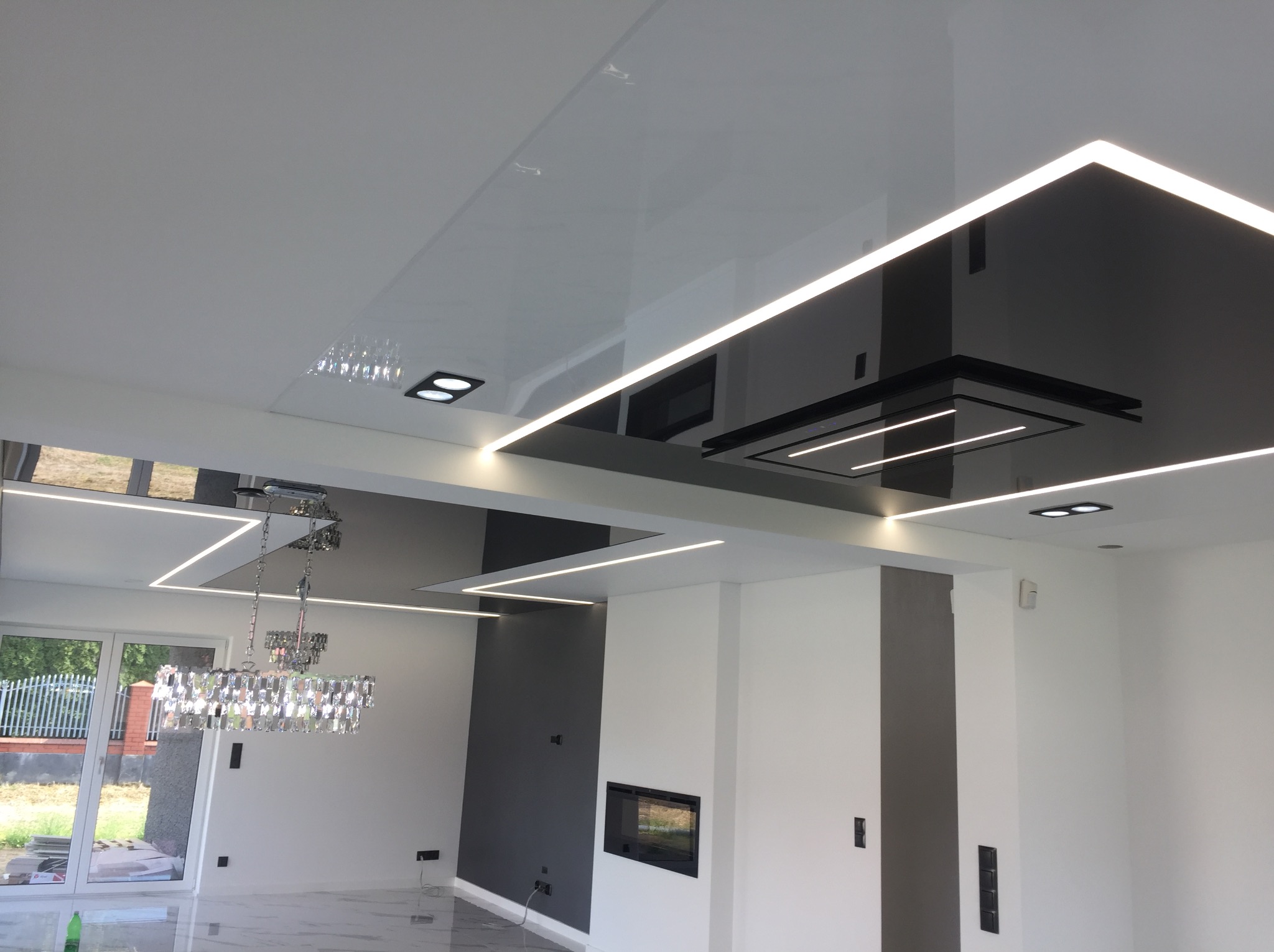 Biało-czarny, błyszczący sufit napinany z oświetleniem LED w nowoczesnym salonie. Widoczny fragment żyrandola i okna. Minimalistyczny design.