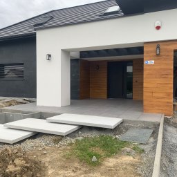 Debruk usługi brukarskie - Nowoczesny dom z betonowymi schodami wejściowymi w trakcie budowy. Widoczne elementy wykończeniowe i przygotowany teren wokół.