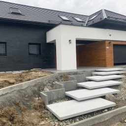Debruk usługi brukarskie - Nowoczesny dom z czarną elewacją i białymi akcentami. Schody z płyt prowadzą do wejścia, teren wokół w trakcie prac ziemnych, widoczne obrzeża i gruz.