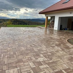 Debruk usługi brukarskie - Nowa, brązowa kostka brukowa ułożona na podjeździe domu z garażem, z widokiem na góry i pochmurne niebo. Widoczne narzędzia w garażu.