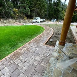 Debruk usługi brukarskie - Kaskadowa ścieżka z kostki brukowej w ogrodzie, obok trawnik i drewniany słup werandy. W tle las i samochód dostawczy. Kostka w odcieniach szarości i brązu.