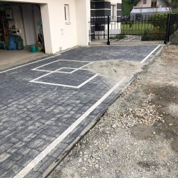 Debruk usługi brukarskie - Szara kostka brukowa z białymi liniami na podjeździe, widok z góry, obok domu z garażem i czarnym ogrodzeniem. Częściowo podsypana piaskiem.