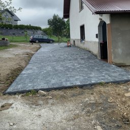 Debruk usługi brukarskie - Ułożona szara kostka brukowa na podjeździe domu jednorodzinnego z widocznym odpływem liniowym i myjką ciśnieniową. W tle zaparkowany samochód osobowy.