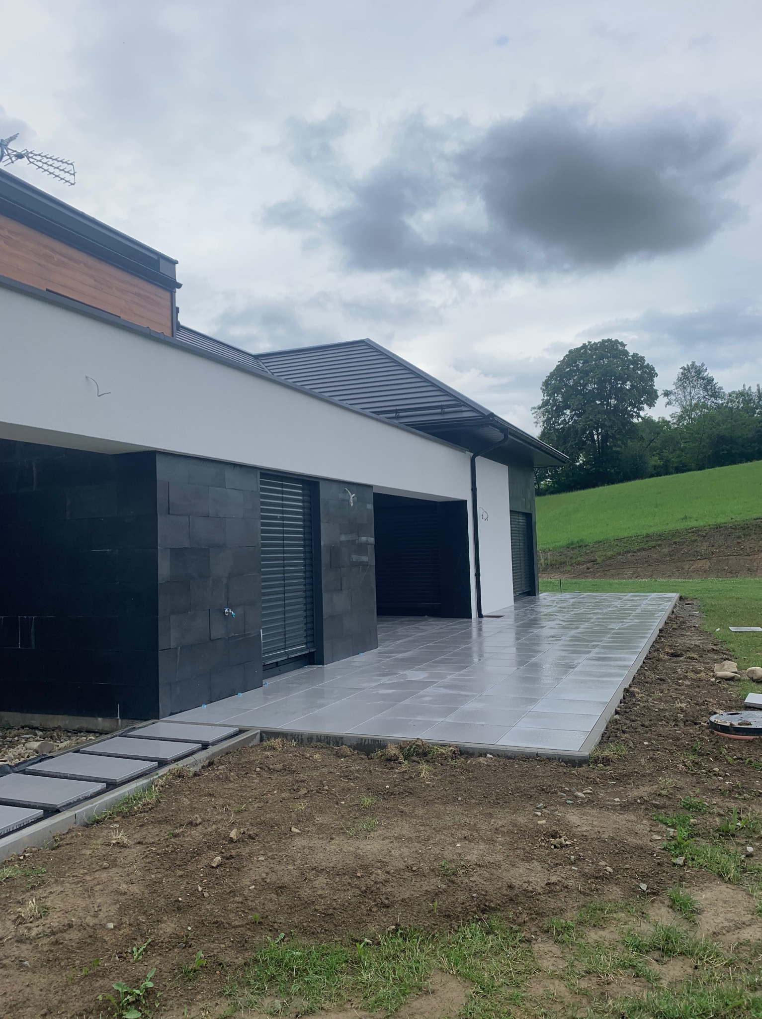 Nowoczesny taras z szarych płyt, przylegający do domu z czarną elewacją. Widoczne przejście z płyt w trawnik, pochmurne niebo w tle.