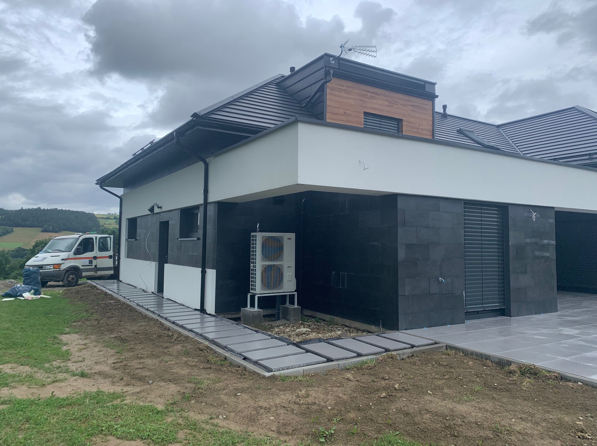 Nowoczesny dom z czarną elewacją i szarym chodnikiem z płyt. Na trawniku stoi biały samochód dostawczy. Pochmurne niebo w tle.