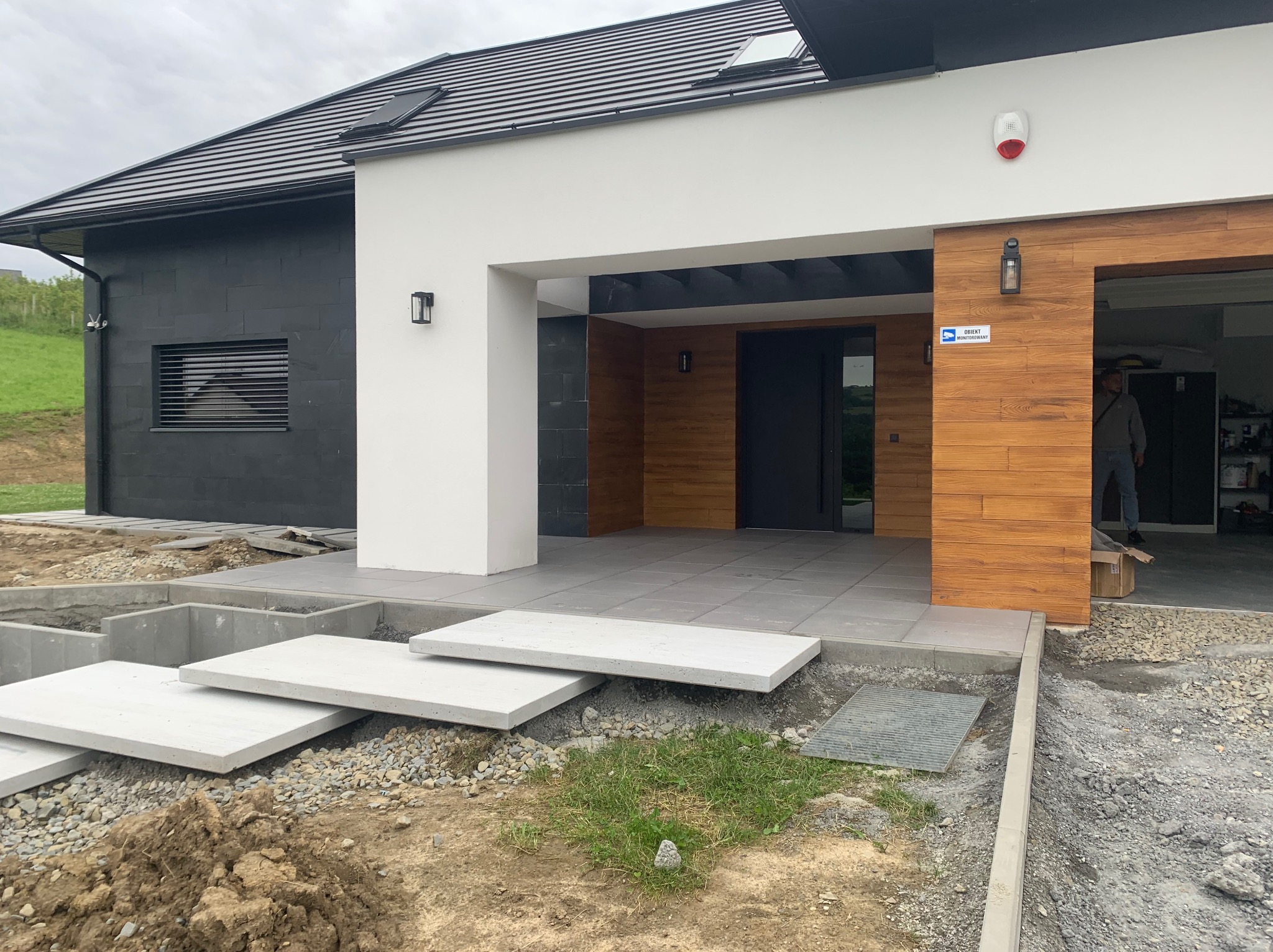 Nowoczesny dom z betonowymi schodami wejściowymi w trakcie budowy. Widoczne elementy wykończeniowe i przygotowany teren wokół.