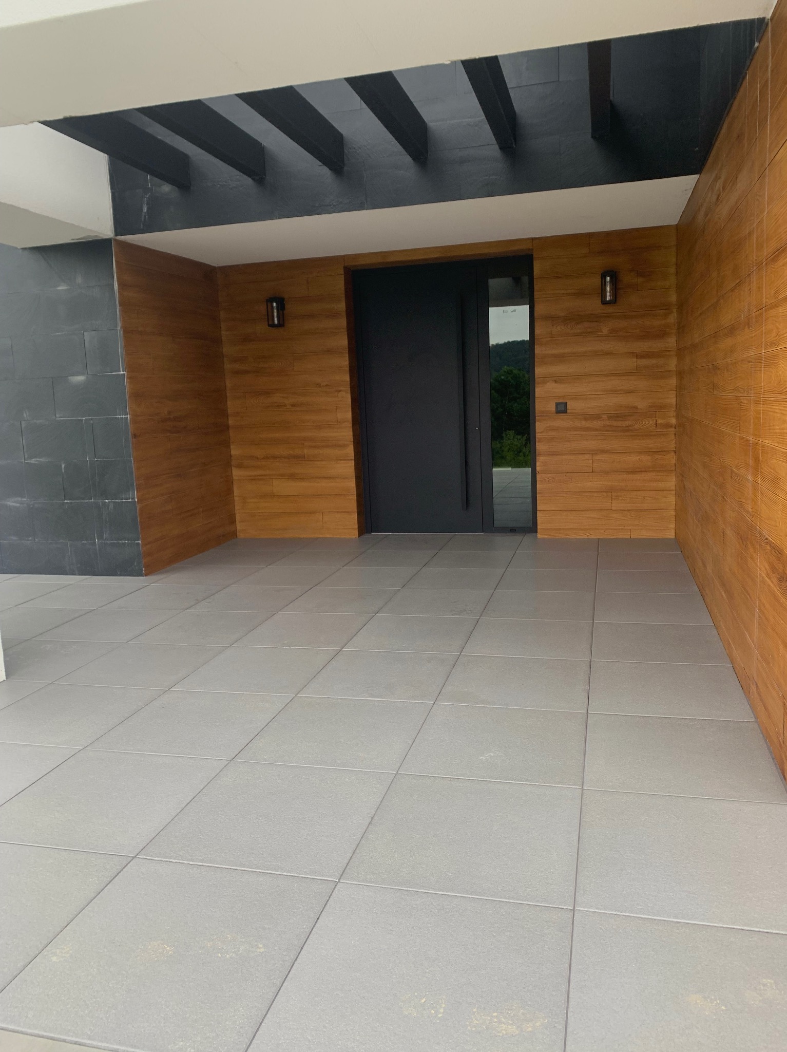 Nowoczesne wejście do domu z szarymi płytami chodnikowymi, drewnianą okładziną ścian i czarną stolarką drzwiową. Minimalistyczny design z elementami drewna.