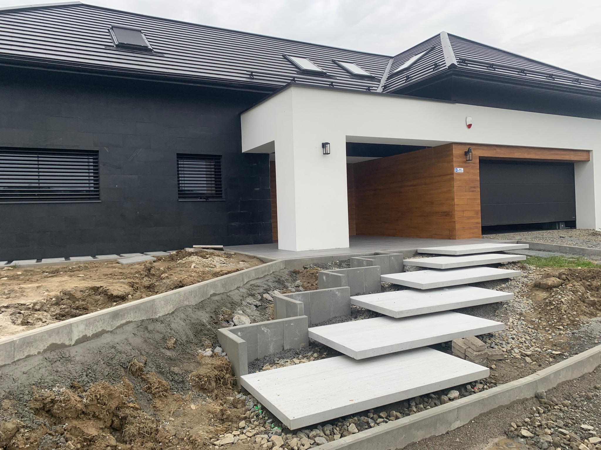 Nowoczesny dom z czarną elewacją i białymi akcentami. Schody z płyt prowadzą do wejścia, teren wokół w trakcie prac ziemnych, widoczne obrzeża i gruz.