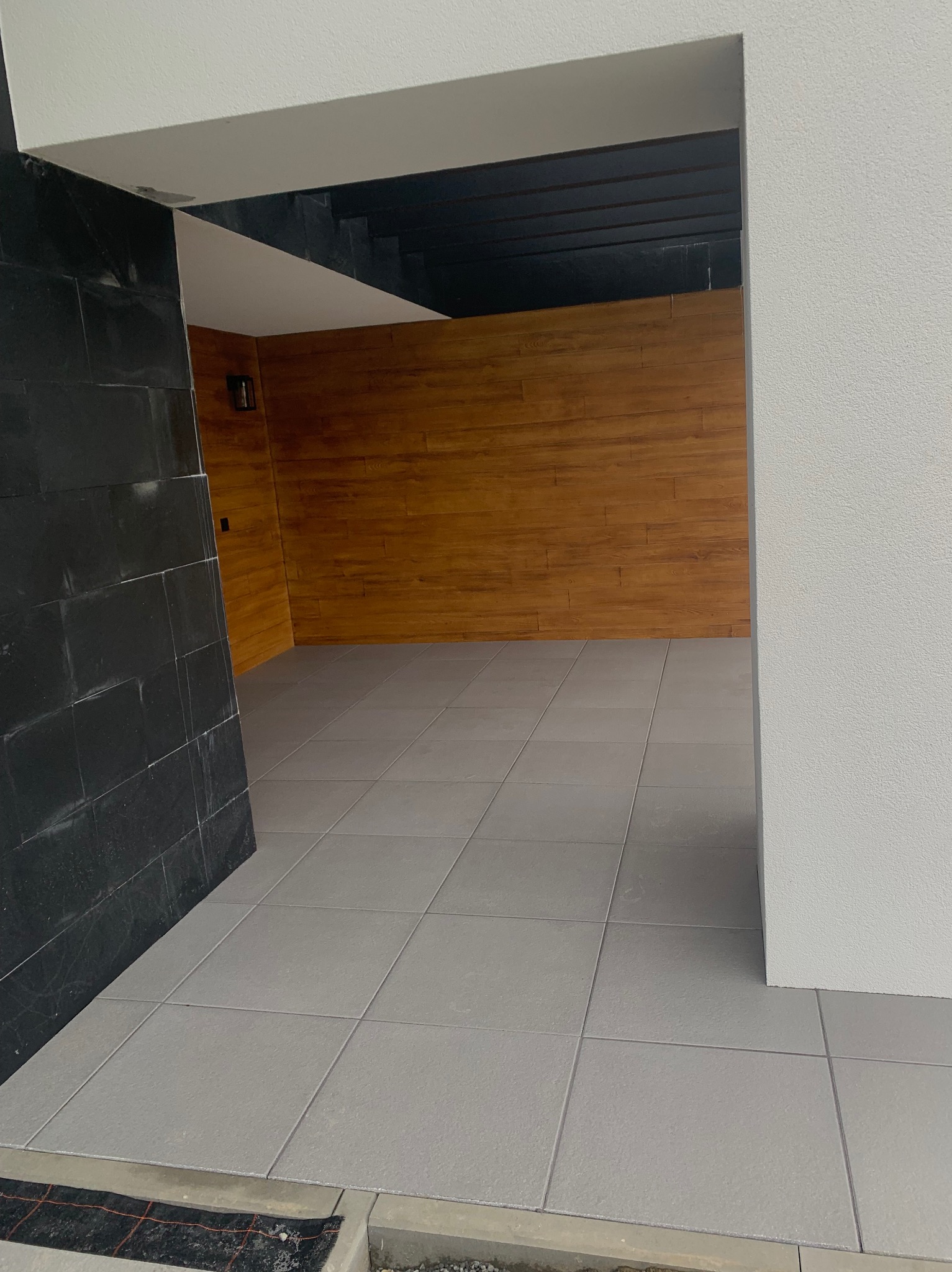 Wykończona, szara podłoga z płytek w przejściu, z widoczną ścianą z drewnianych paneli i czarną, kamienną ścianą po lewej stronie. Minimalistyczny design.