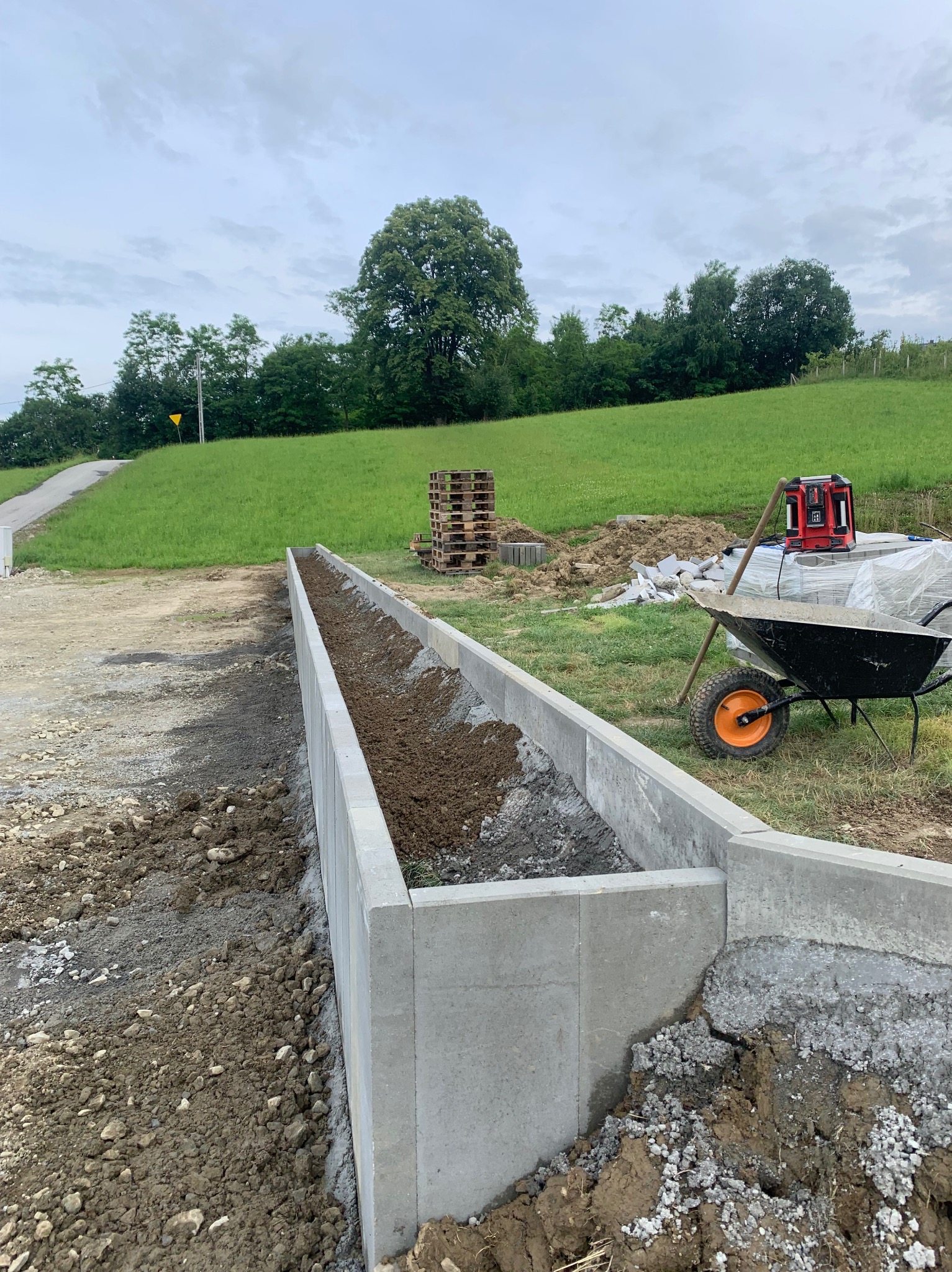 Betonowe obrzeże w trakcie budowy w Krakowie, widoczna taczka i laserowy niwelator. Obrzeże wypełnione ziemią, w tle zielone wzgórza i drzewa.