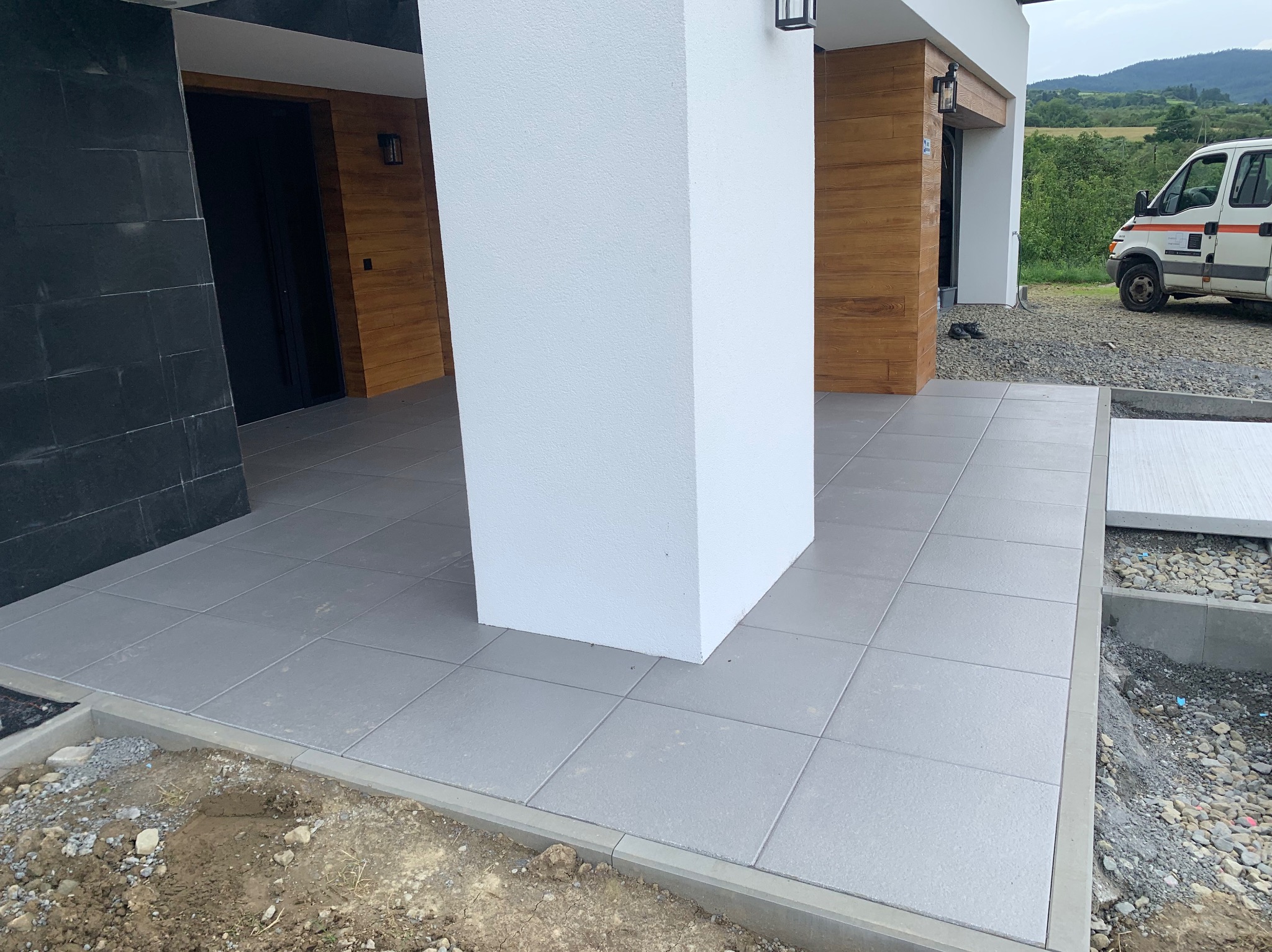 Nowoczesny taras z szarych płyt, wykończony betonową obwódką. Widoczny fragment elewacji budynku z drewnianymi panelami i kolumna. W tle krajobraz.