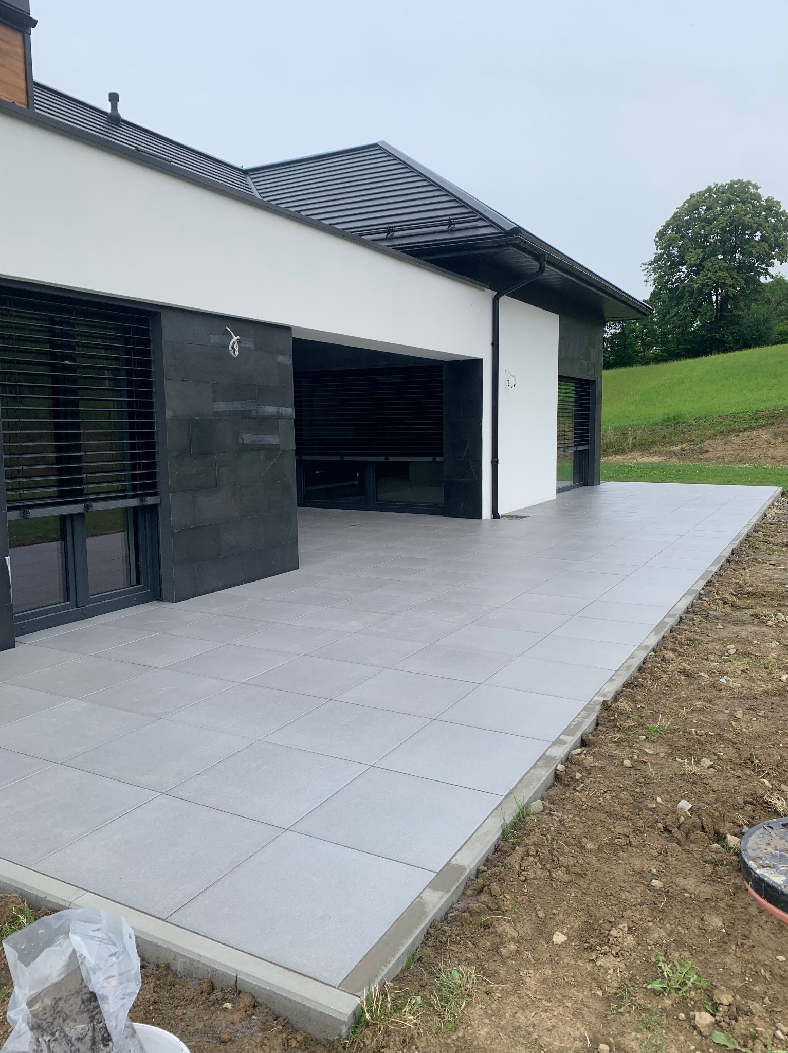 Nowoczesny taras z szarych płyt, przylegający do domu z czarno-białą elewacją. Widoczne zielone wzgórze w tle i fragment niezabudowanego terenu.