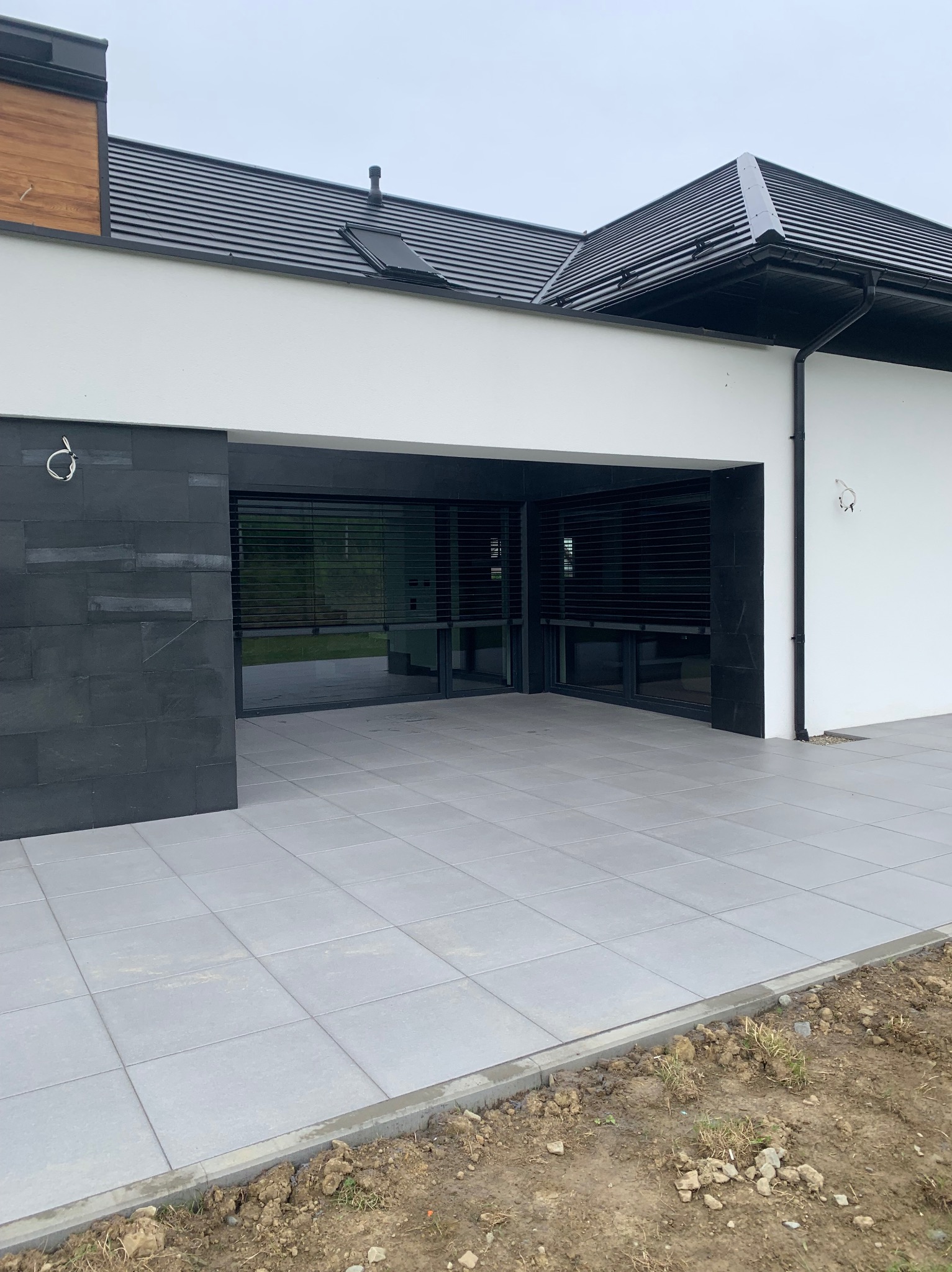 Nowoczesny taras z szarych płyt, przylegający do budynku z czarnymi elementami elewacji i dachem. Widoczne niedokończone obrzeża tarasu.