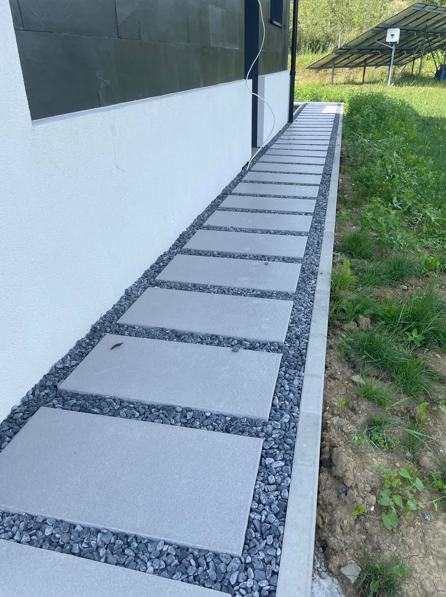 Nowoczesna ścieżka z płyt betonowych i grysu przy białej ścianie budynku. W tle panele słoneczne i zieleń. Minimalistyczny design.