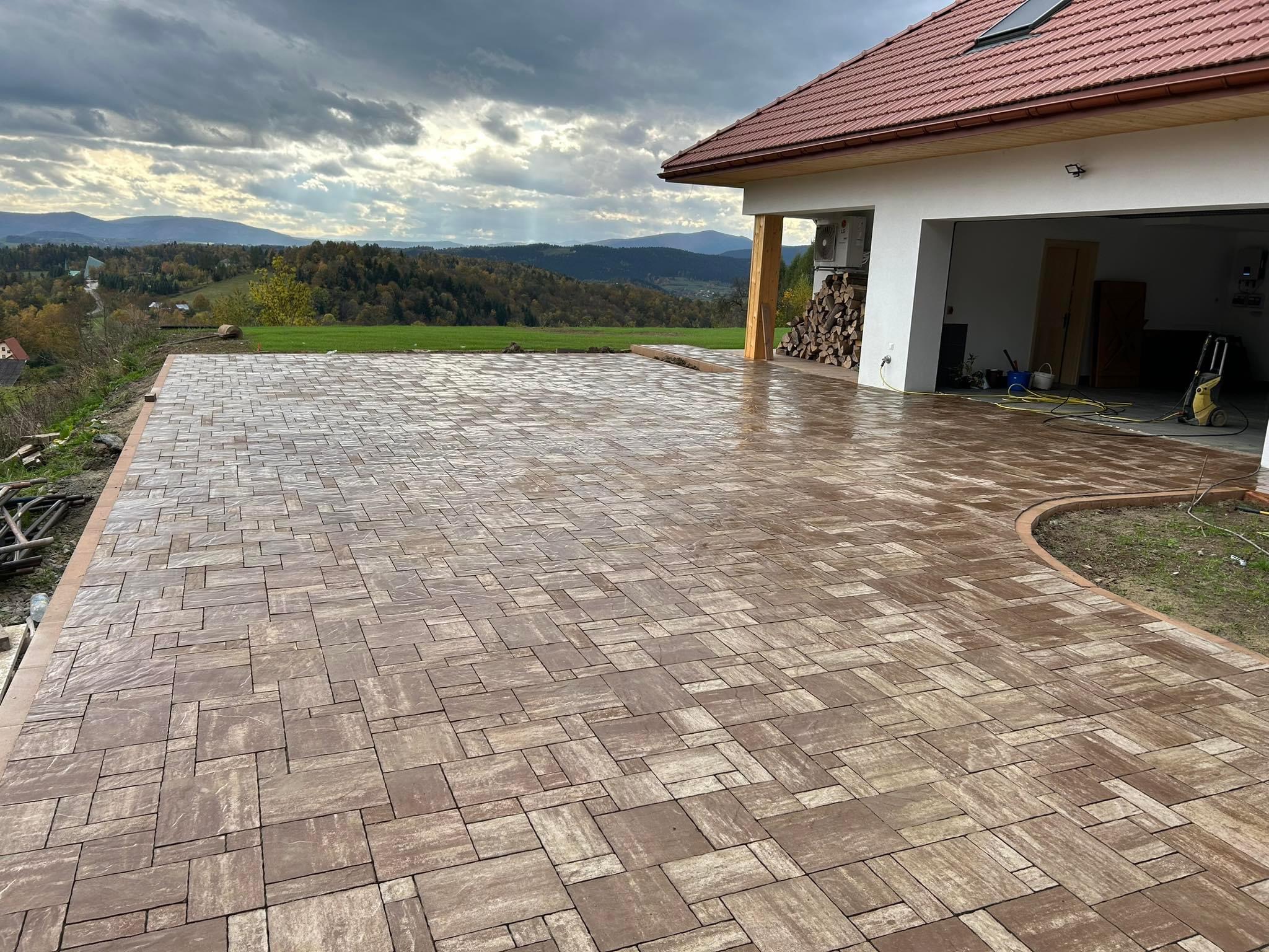 Nowa, brązowa kostka brukowa ułożona na podjeździe domu z garażem, z widokiem na góry i pochmurne niebo. Widoczne narzędzia w garażu.