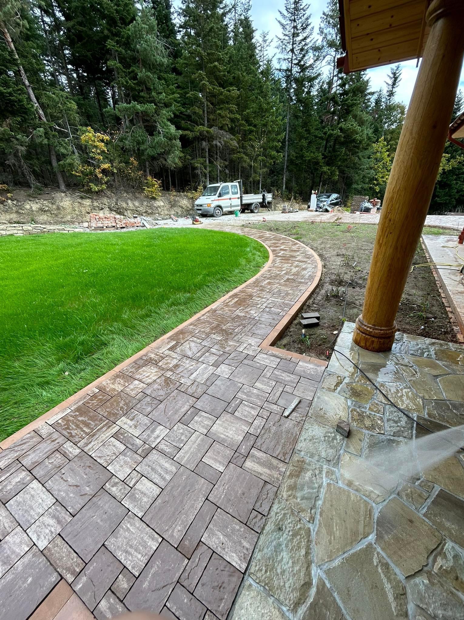 Kaskadowa ścieżka z kostki brukowej w ogrodzie, obok trawnik i drewniany słup werandy. W tle las i samochód dostawczy. Kostka w odcieniach szarości i brązu.