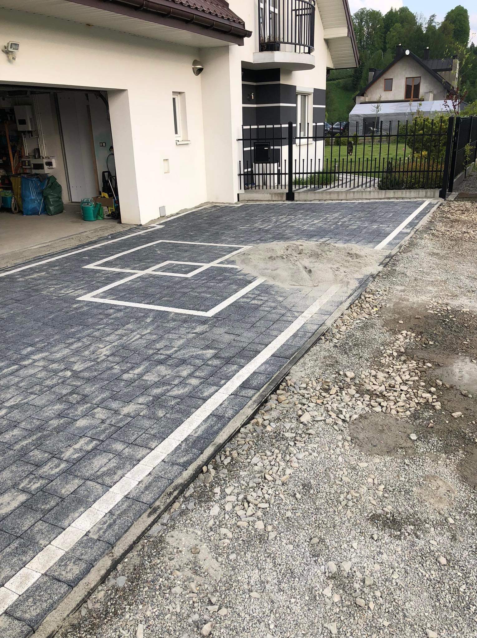 Szara kostka brukowa z białymi liniami na podjeździe, widok z góry, obok domu z garażem i czarnym ogrodzeniem. Częściowo podsypana piaskiem.