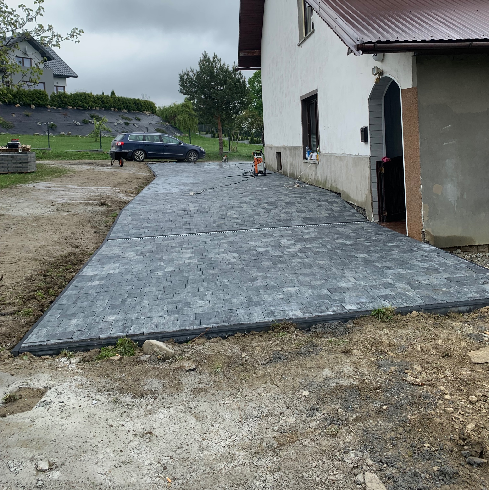 Ułożona szara kostka brukowa na podjeździe domu jednorodzinnego z widocznym odpływem liniowym i myjką ciśnieniową. W tle zaparkowany samochód osobowy.