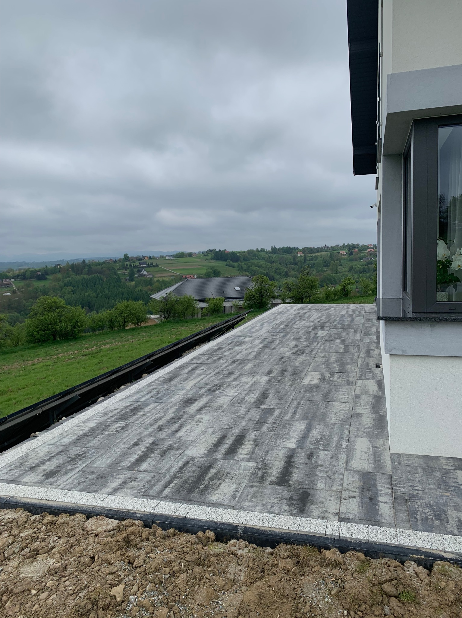 Ułożona kostka brukowa w odcieniach szarości na tarasie z widokiem na zielone wzgórza. Widoczny fragment budynku z oknem. Brzeg tarasu wykończony drobną kostką.