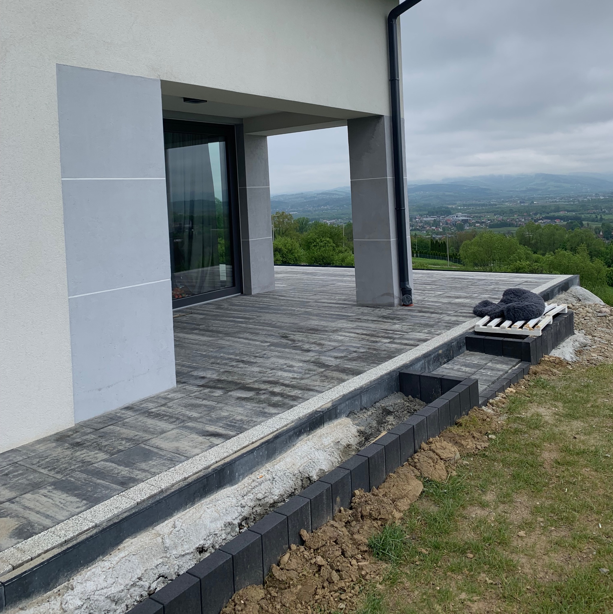 Nowoczesny taras z szarej kostki brukowej, obrzeża z ciemnej kostki. Widok na góry w tle. Częściowo ukończone prace.