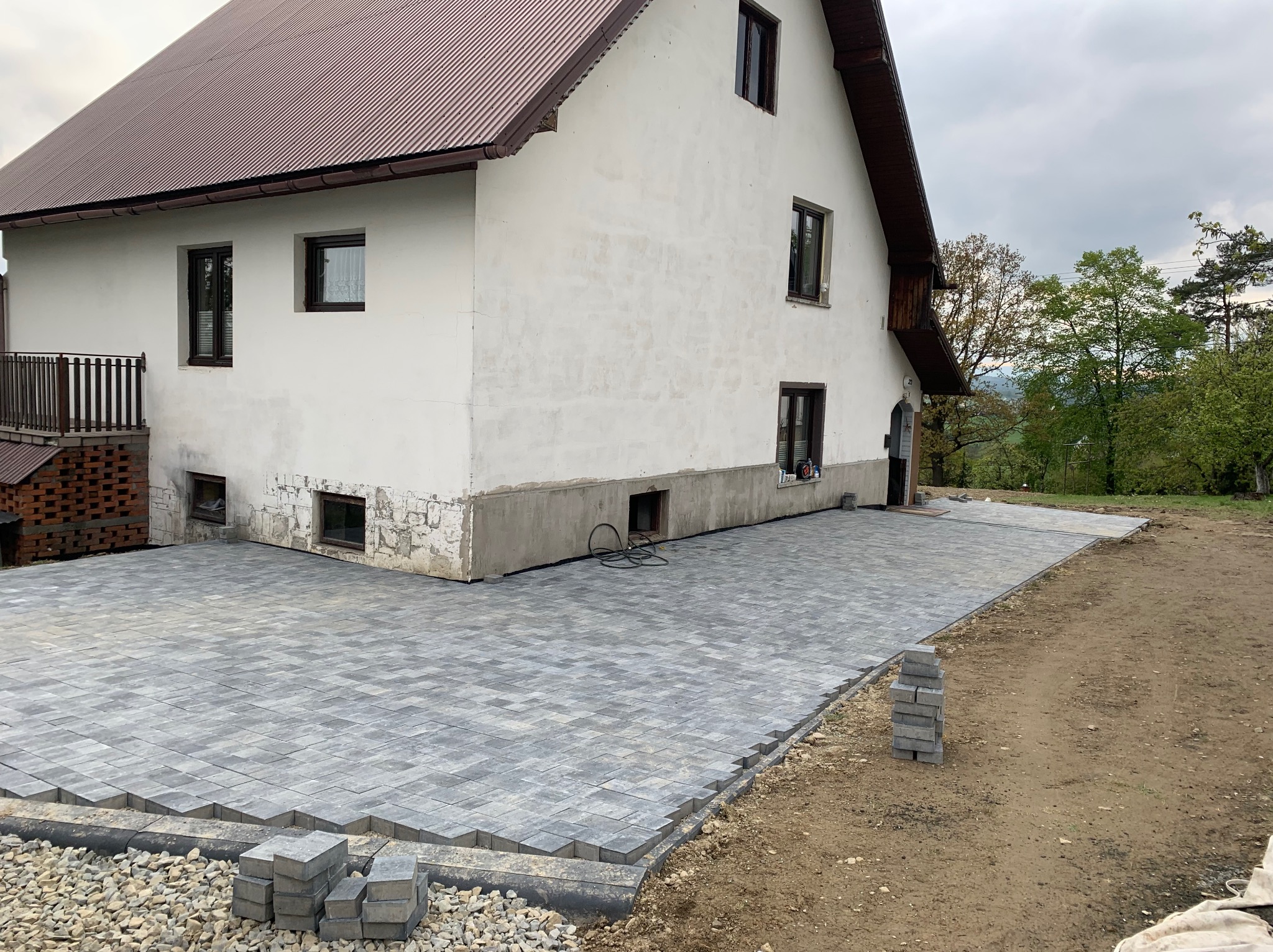 Szara kostka brukowa ułożona przed białym domem z brązowym dachem. Część kostki jeszcze nieułożona, leży w stosie. Widoczne obrzeża i kamienie.