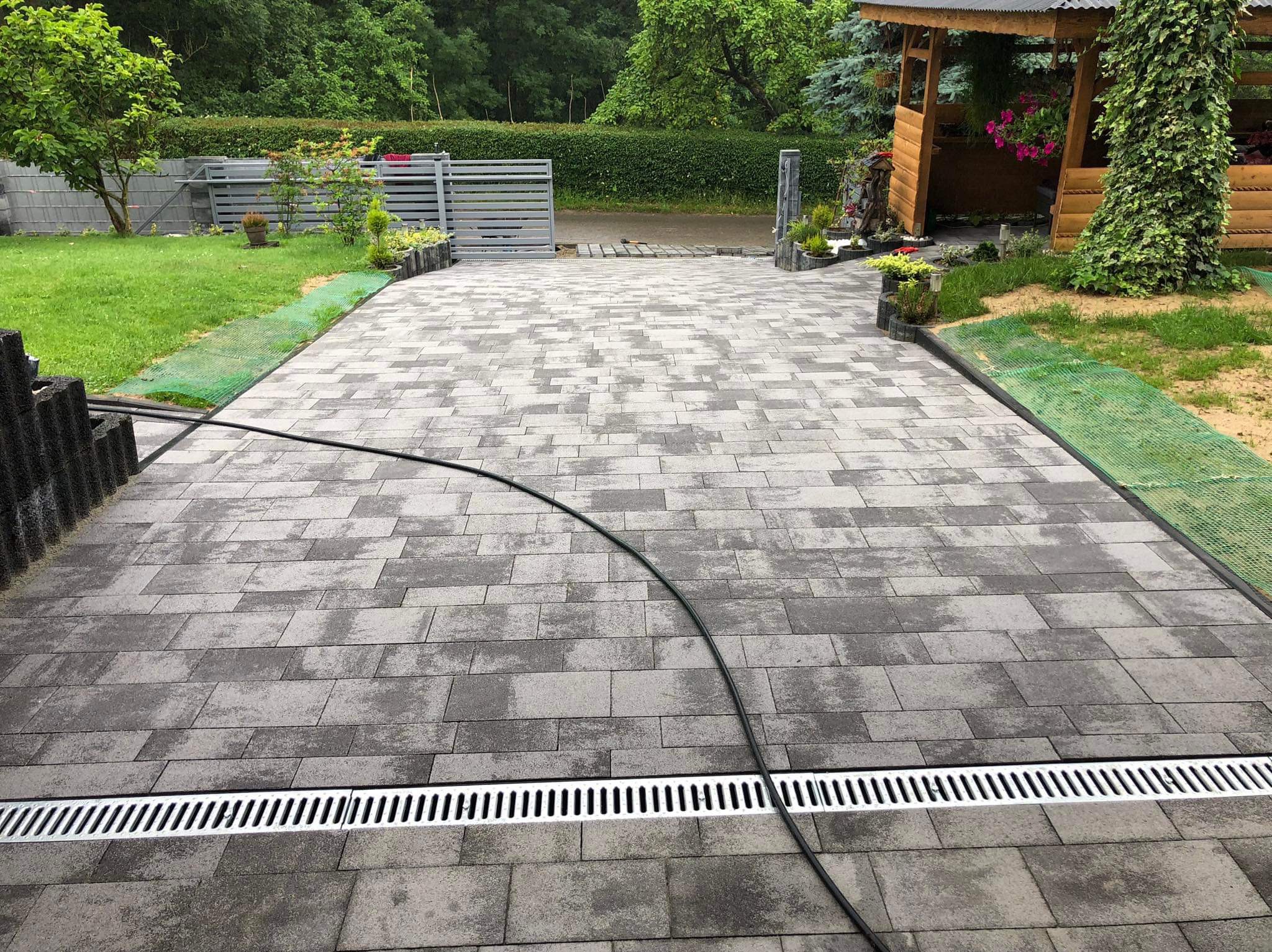 Szara kostka brukowa ułożona na podjeździe z odpływem liniowym, widok na bramę i altanę w ogrodzie. Estetyczne wykończenie posesji.