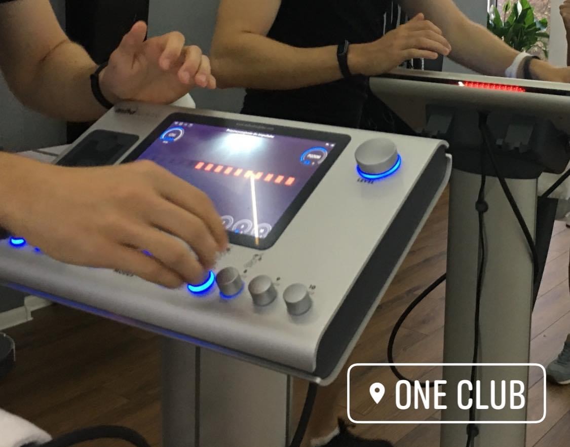 Obsługa panelu sterowania urządzenia EMS z podświetlanymi przyciskami, widoczne dłonie osoby obsługującej i fragmenty ciał osób ćwiczących. Studio treningowe ONE CLUB.