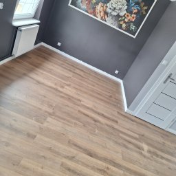 Xyz - Jasne panele podłogowe ułożone w pokoju z szarą ścianą, oknem dachowym i obrazem w kwiaty. Widok na fragment drzwi.