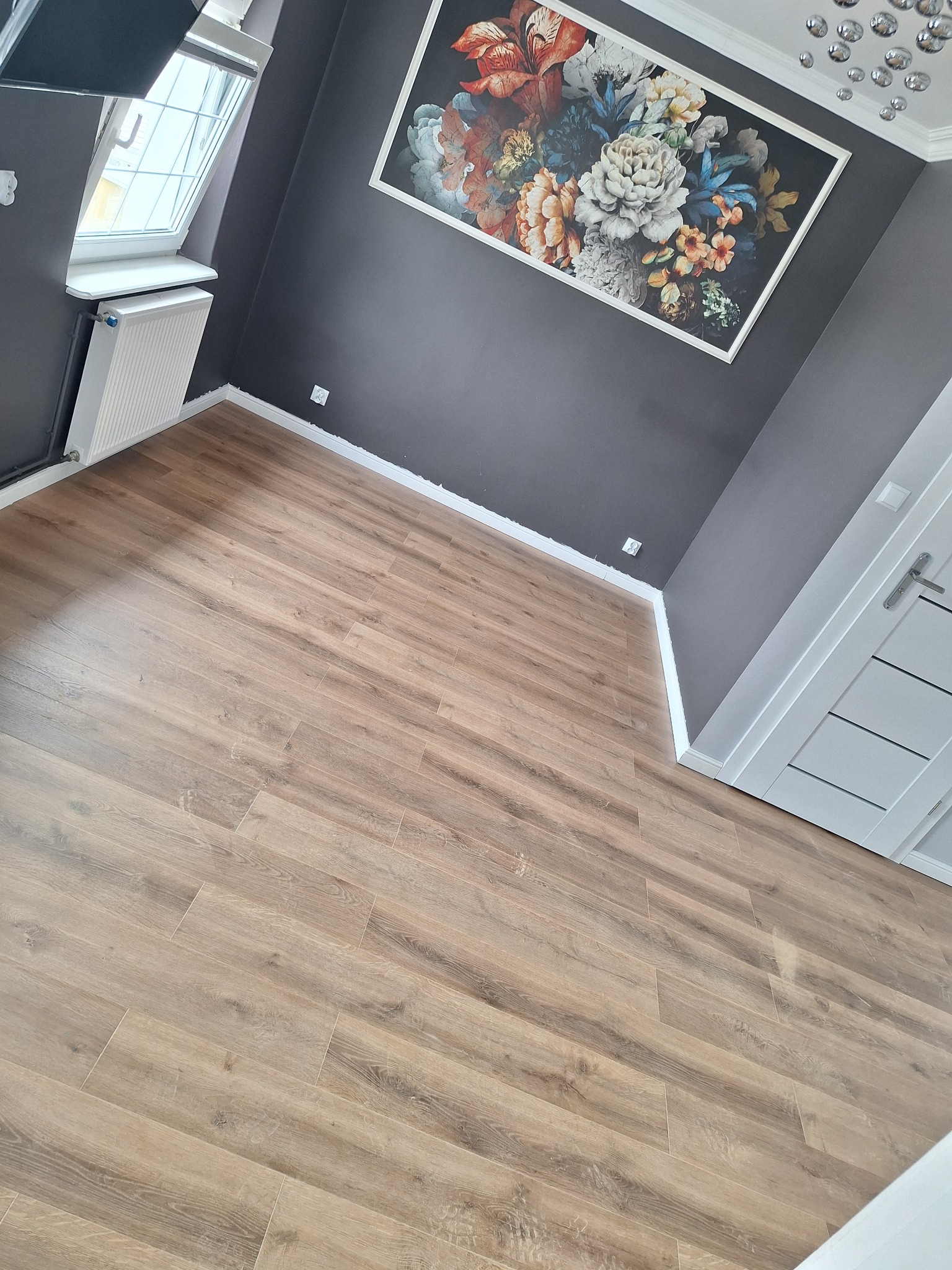 Jasne panele podłogowe ułożone w pokoju z szarą ścianą, oknem dachowym i obrazem w kwiaty. Widok na fragment drzwi.
