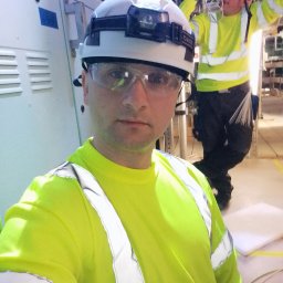 Atlarion Technika - Elektryk w kasku i okularach ochronnych, w jaskrawej kamizelce, na tle szafy sterowniczej i drugiego elektryka przy instalacji. Praca w pomieszczeniu technicznym.