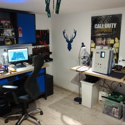 Atlarion Technika - Warsztat elektronika z biurkiem, komputerem z projektem płytki PCB, narzędziami, szafkami w kolorze niebieskim i plakatem gry Assassin's Creed Origins.