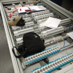 Atlarion Technika - Rozdzielnica elektryczna w trakcie montażu, widoczne bezpieczniki, kable, narzędzia i dokumentacja. Elementy ułożone wewnątrz metalowej obudowy.