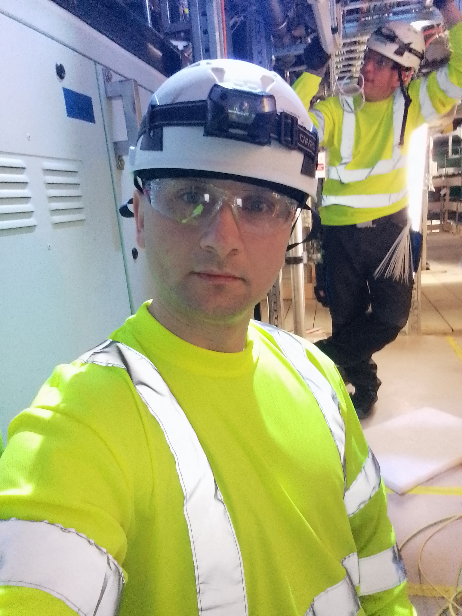 Elektryk w kasku i okularach ochronnych, w jaskrawej kamizelce, na tle szafy sterowniczej i drugiego elektryka przy instalacji. Praca w pomieszczeniu technicznym.