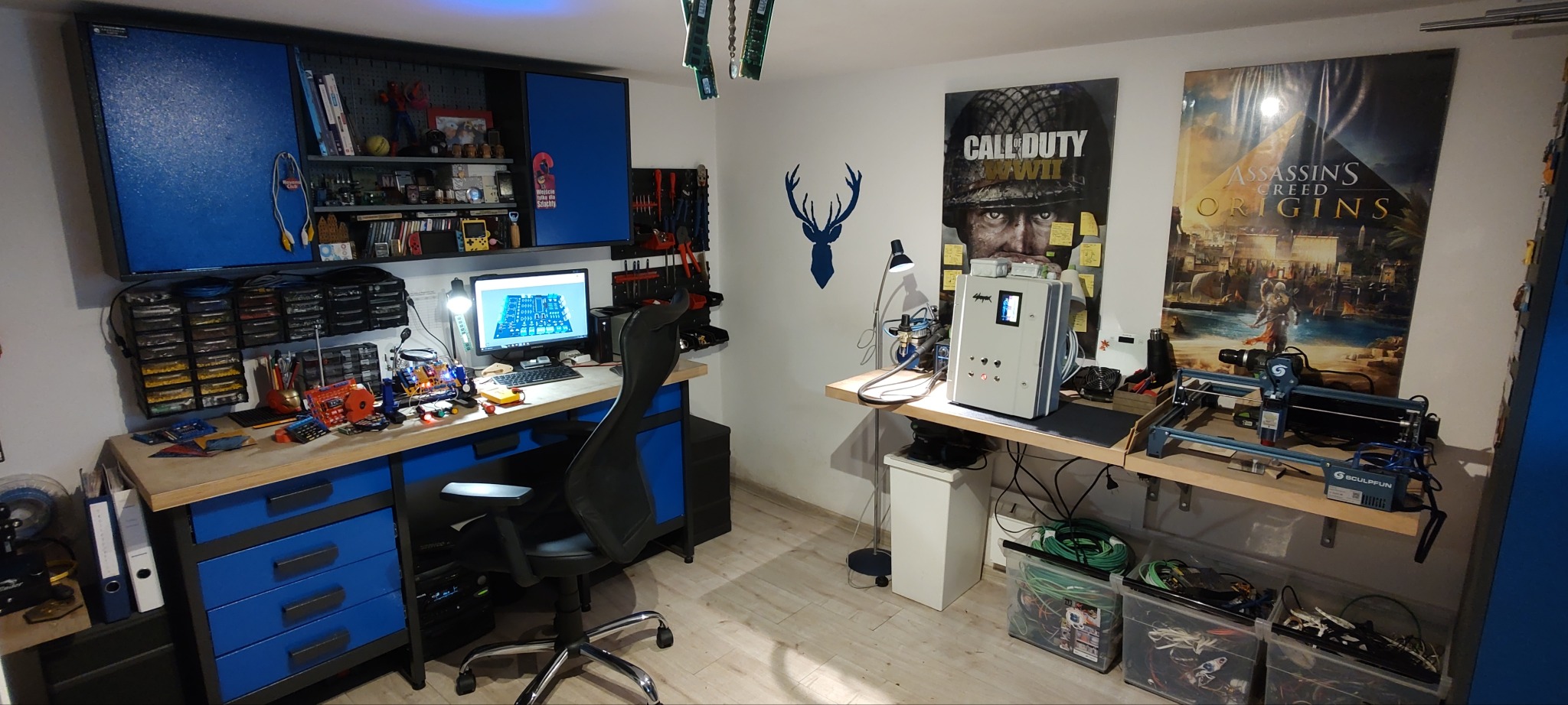 Warsztat elektronika z biurkiem, komputerem z projektem płytki PCB, narzędziami, szafkami w kolorze niebieskim i plakatem gry Assassin's Creed Origins.