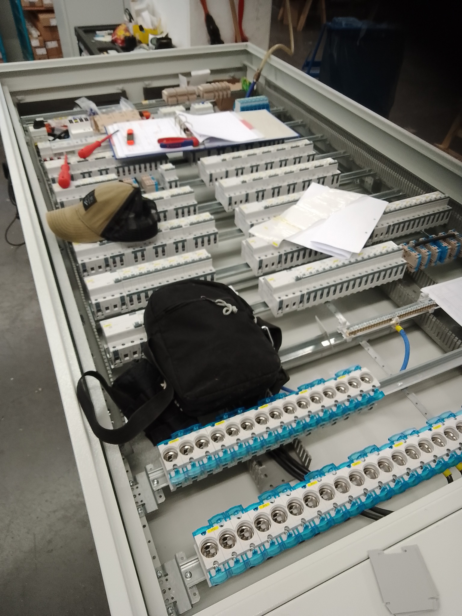 Rozdzielnica elektryczna w trakcie montażu, widoczne bezpieczniki, kable, narzędzia i dokumentacja. Elementy ułożone wewnątrz metalowej obudowy.