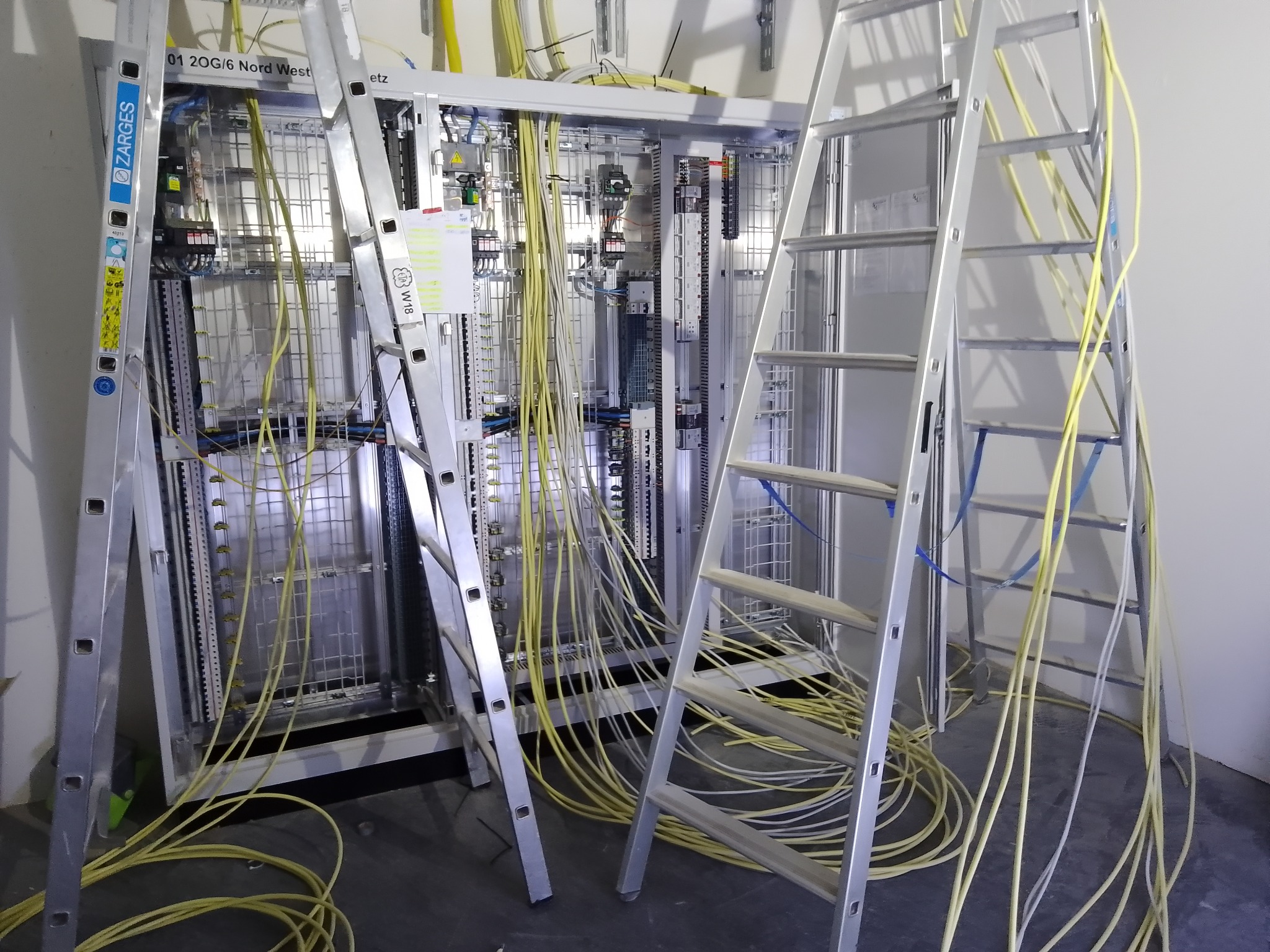 Rozbudowana szafa sterownicza z licznymi przewodami w kolorze żółtym, dostęp do niej zapewniają dwie aluminiowe drabiny. Widoczne elementy instalacji elektrycznej.