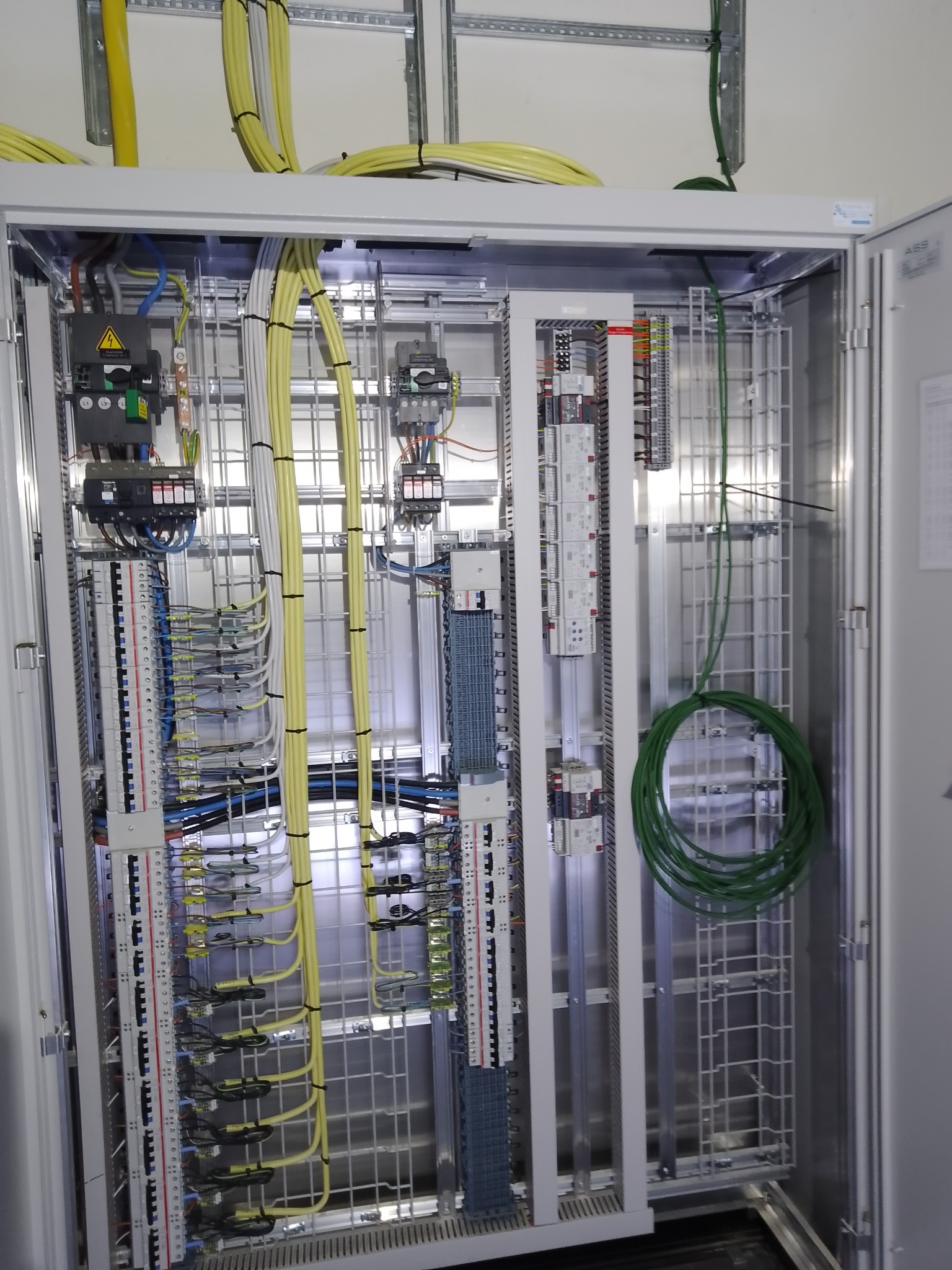 Otwarta szafa sterownicza z precyzyjnie ułożonymi kablami w kolorach żółtym, zielonym i niebieskim, zamocowanymi uchwytami. Widoczne zabezpieczenia i moduły.
