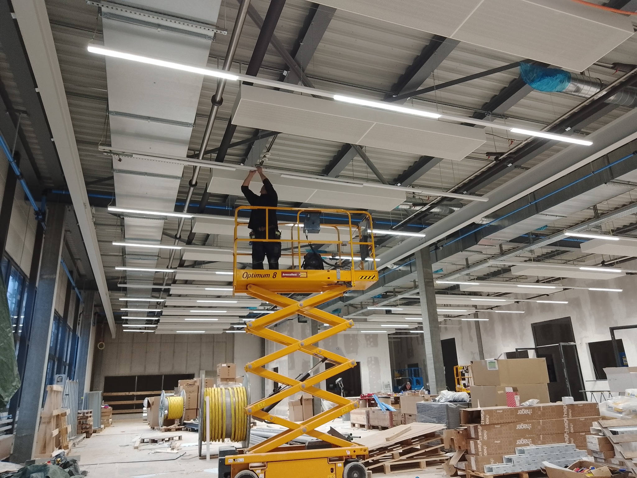 Montaż paneli sufitowych w hali przemysłowej z użyciem podnośnika nożycowego. Widoczne elementy instalacji oświetleniowej i wentylacyjnej.