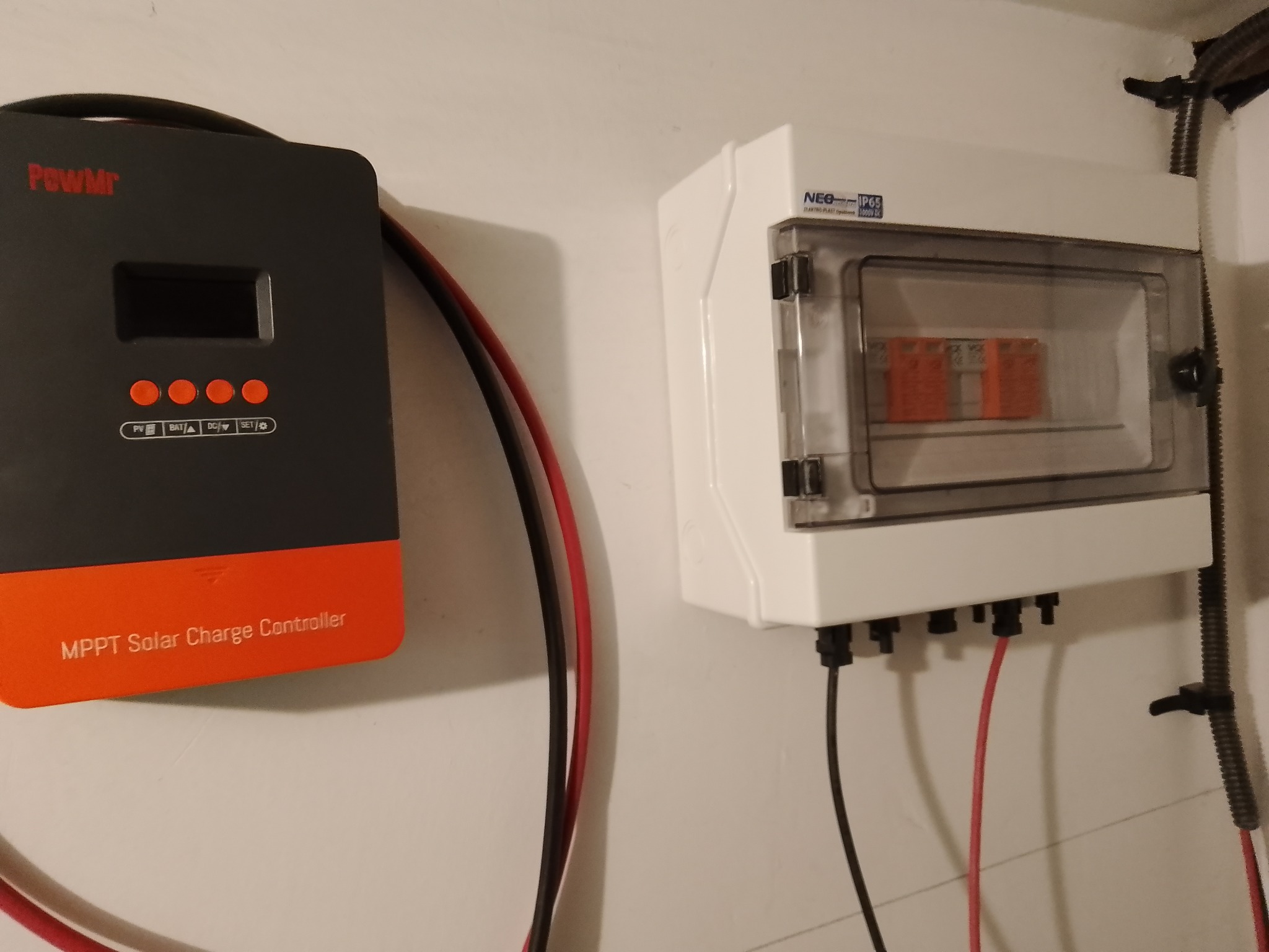 Szaro-pomarańczowy regulator ładowania solarnego PowMr obok białej skrzynki elektrycznej NEO IP65 z widocznymi bezpiecznikami, zamontowane na białej ścianie.