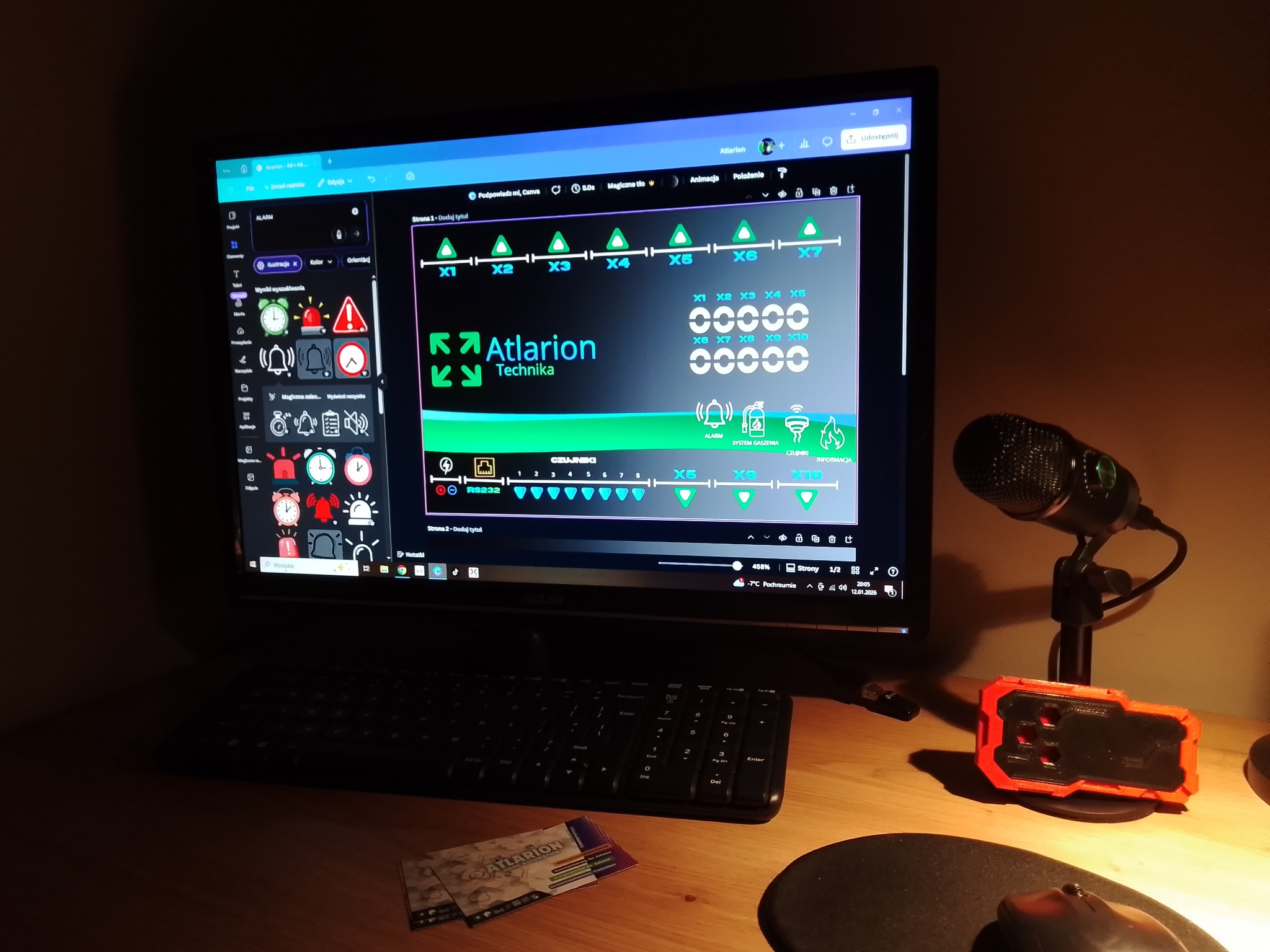 Ekran monitora z otwartym projektem systemu alarmowego 'Atlarion Technika', widoczne ikony czujników i schemat. Obok mikrofon i urządzenie pomiarowe na biurku.