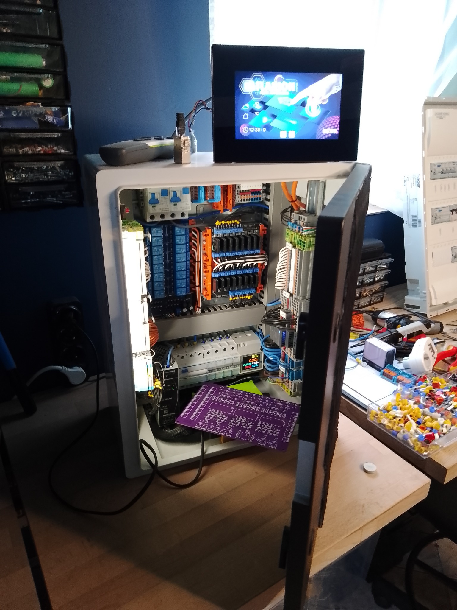 Otwarta szafa sterownicza z widocznymi elementami elektrycznymi, przewodami i ekranem dotykowym na górze. Widoczna płytka PCB i elementy elektroniczne na stole.