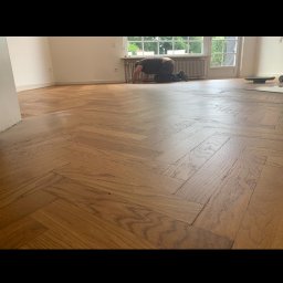 M&L Parkiet - Parkiet ułożony w jodełkę, świeżo po renowacji. Widoczny pracownik w trakcie wykańczania podłogi przy oknie. Jasne wnętrze, naturalne światło.